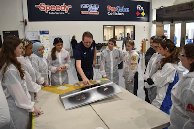 Instructor demonstrates automotive repair technique to students in white lab coats at a training facility.