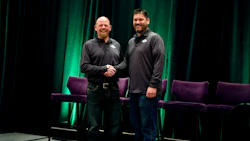 Two men shake hands on stage during a professional event with green lighting and purple seating
