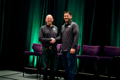 Two men shake hands on stage during a professional event with green lighting and purple seating