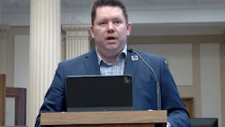 Man in blue blazer speaking at wooden podium with microphone in formal indoor setting