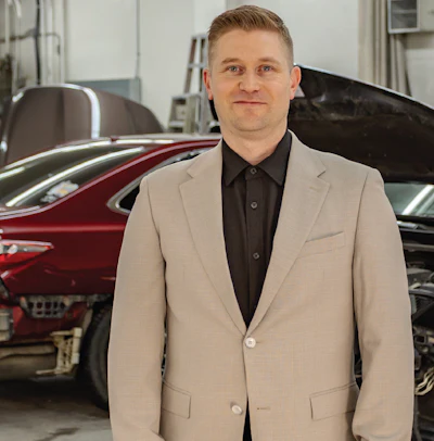 Professional man in beige blazer and black shirt standing in car showroom with vehicles in background