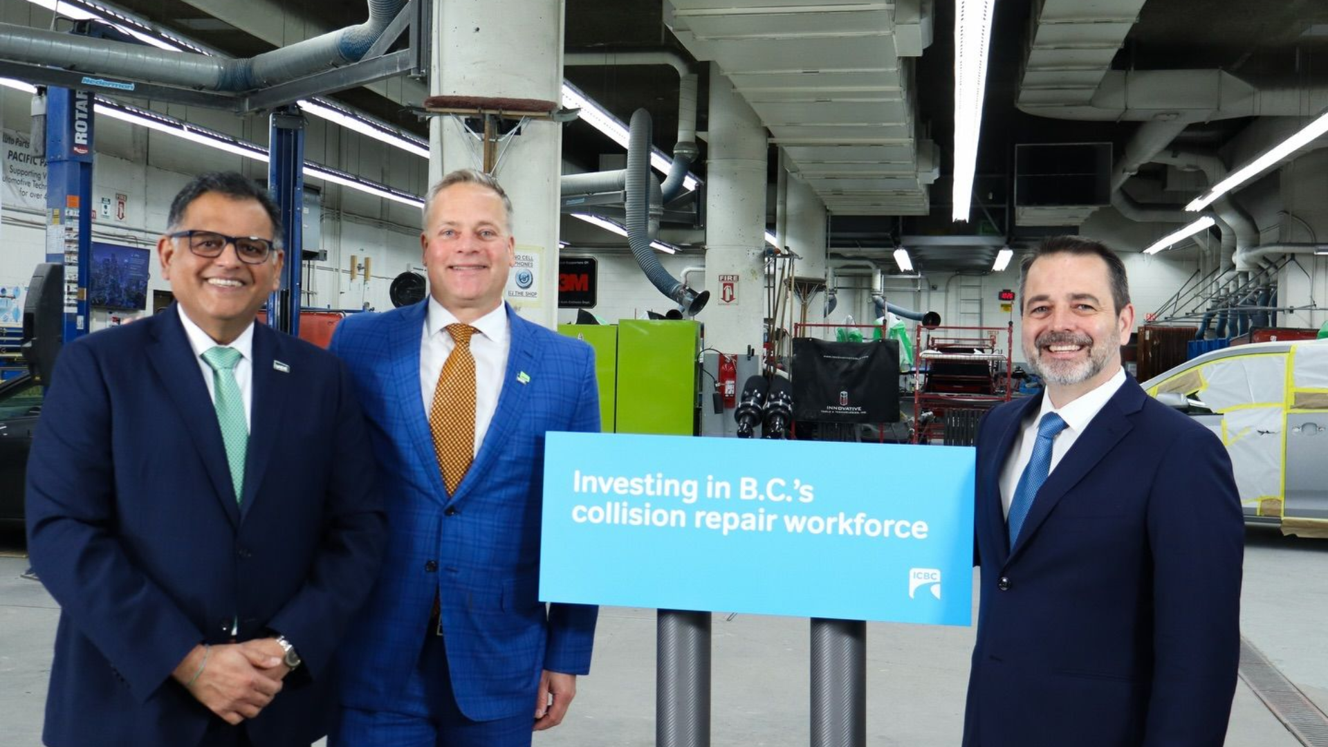Three business professionals holding blue banner promoting investment in B.C.'s collision repair workforce at industrial facility