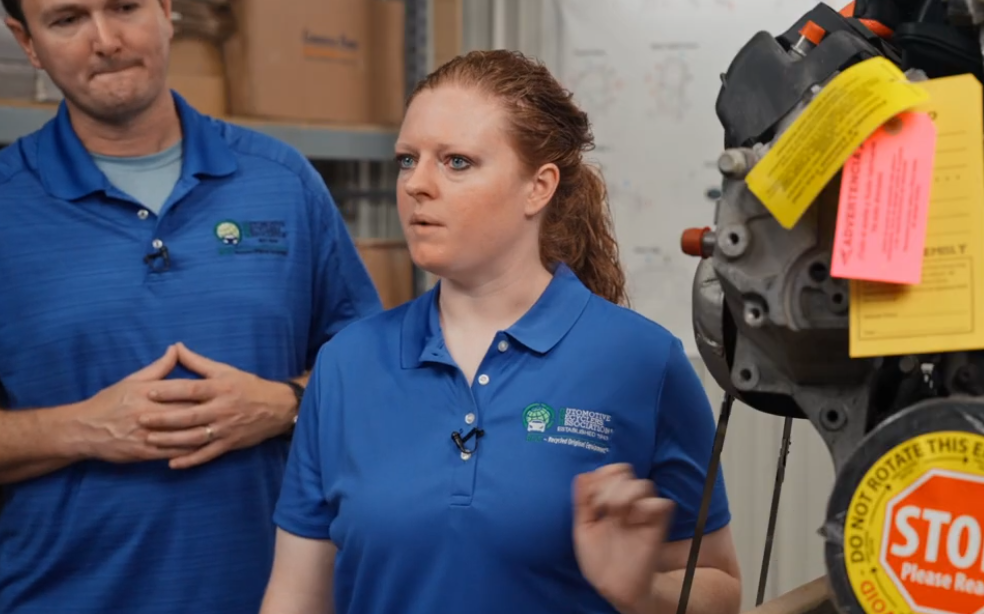 Two employees in blue branded polo shirts conduct a training demonstration in a warehouse facility with equipment visible in background.