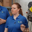 Two employees in blue branded polo shirts conduct a training demonstration in a warehouse facility with equipment visible in background.