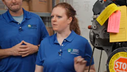 Two employees in blue branded polo shirts conduct a training demonstration in a warehouse facility with equipment visible in background.