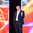 Professional man in dark blazer speaking on stage with abstract red gradient display screens behind him