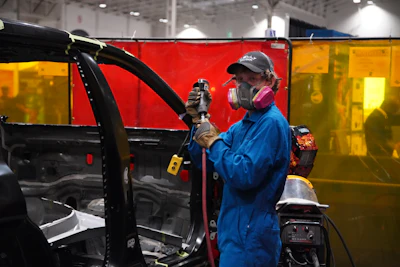 Welder in blue coveralls and protective gear working on red vehicle door with welding torch in industrial workshop