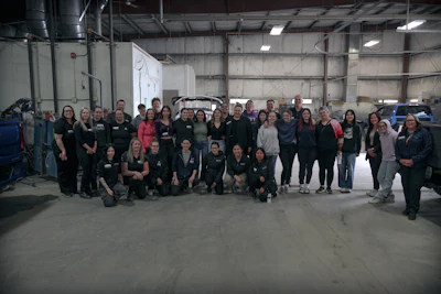 Large diverse team group photo in industrial warehouse setting with vehicles in background