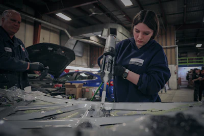 Female mechanic using pneumatic drill on vehicle in professional automotive workshop with colleague and cars in background