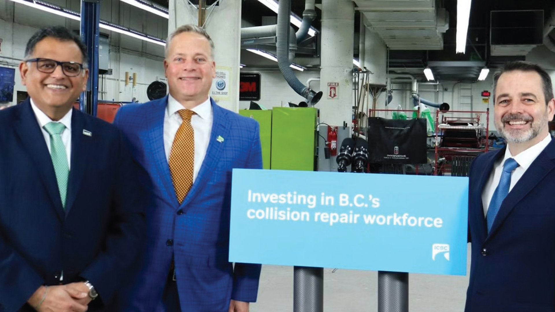 Three business professionals hold a light blue sign announcing investment in B.C.'s collision repair workforce at an auto repair facility.