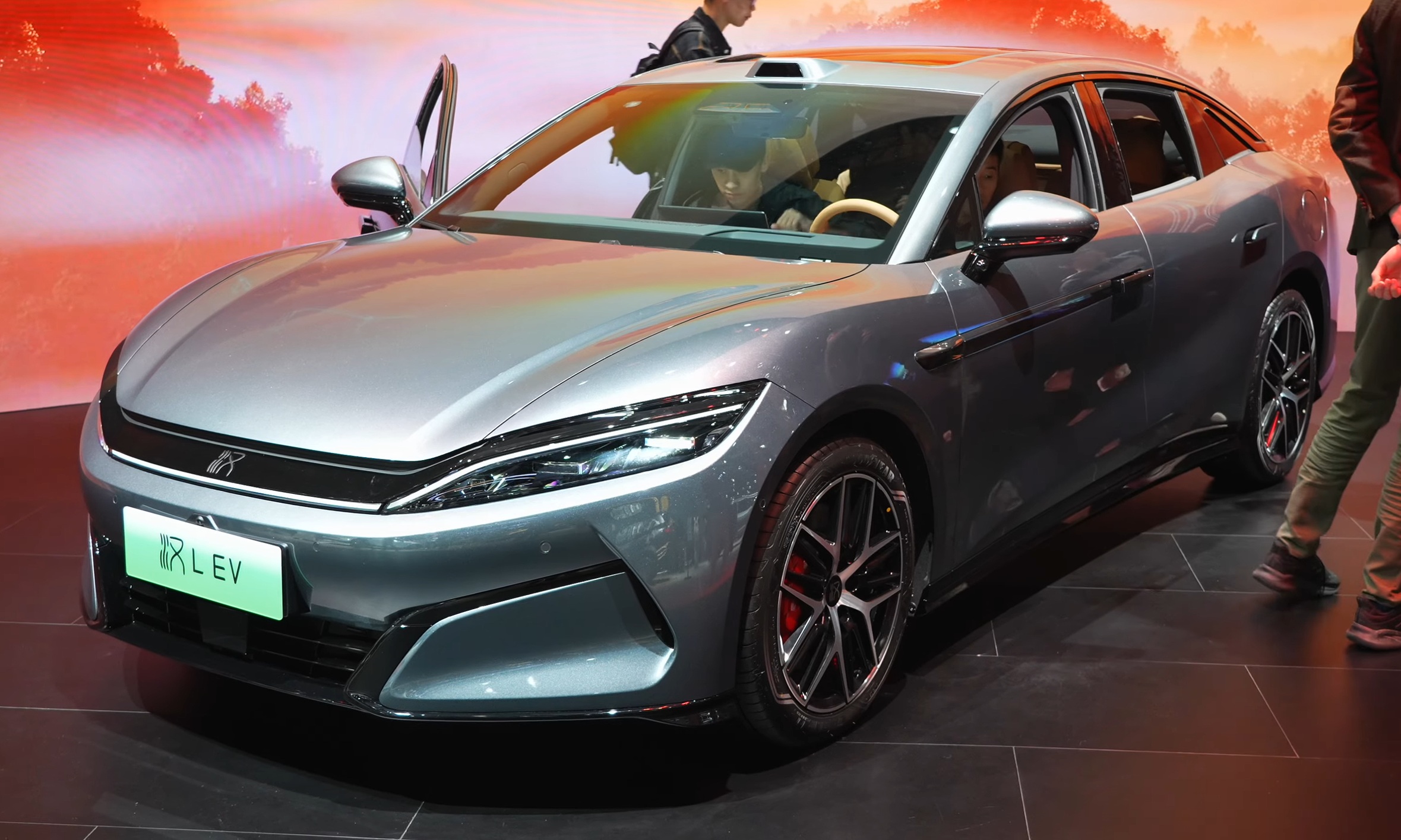 Silver BYD electric sedan on display at automotive exhibition with dramatic orange sunset backdrop