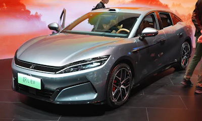 Silver BYD electric sedan on display at automotive exhibition with dramatic orange sunset backdrop