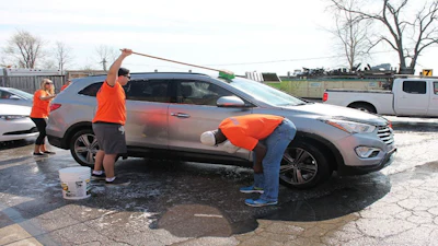 Volunteers in orange shirts washing a gray SUV at an outdoor car wash event