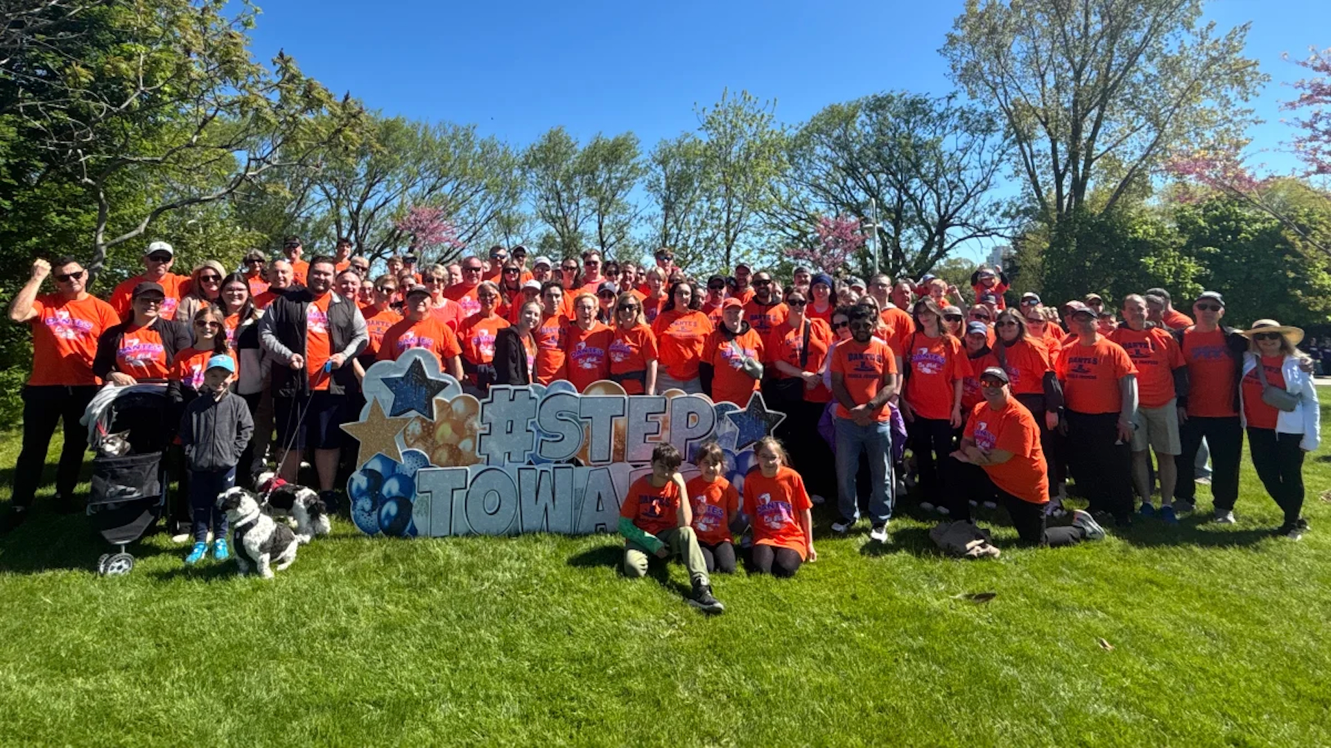 Large group of people in orange shirts gathered in a park holding a blue and white 'FIRST TOWN' sign
