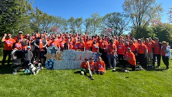Large group of people in orange shirts gathered in a park holding a blue and white 'FIRST TOWN' sign