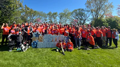 Large group of people in orange shirts gathered in a park holding a blue and white 'FIRST TOWN' sign