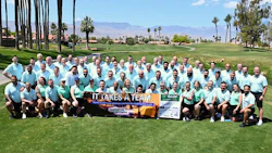 Large team group photo on golf course with 60+ people in light blue and green shirts holding charity banners
