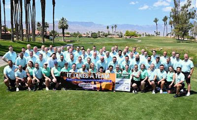 Large team group photo on golf course with 60+ people in light blue and green shirts holding charity banners