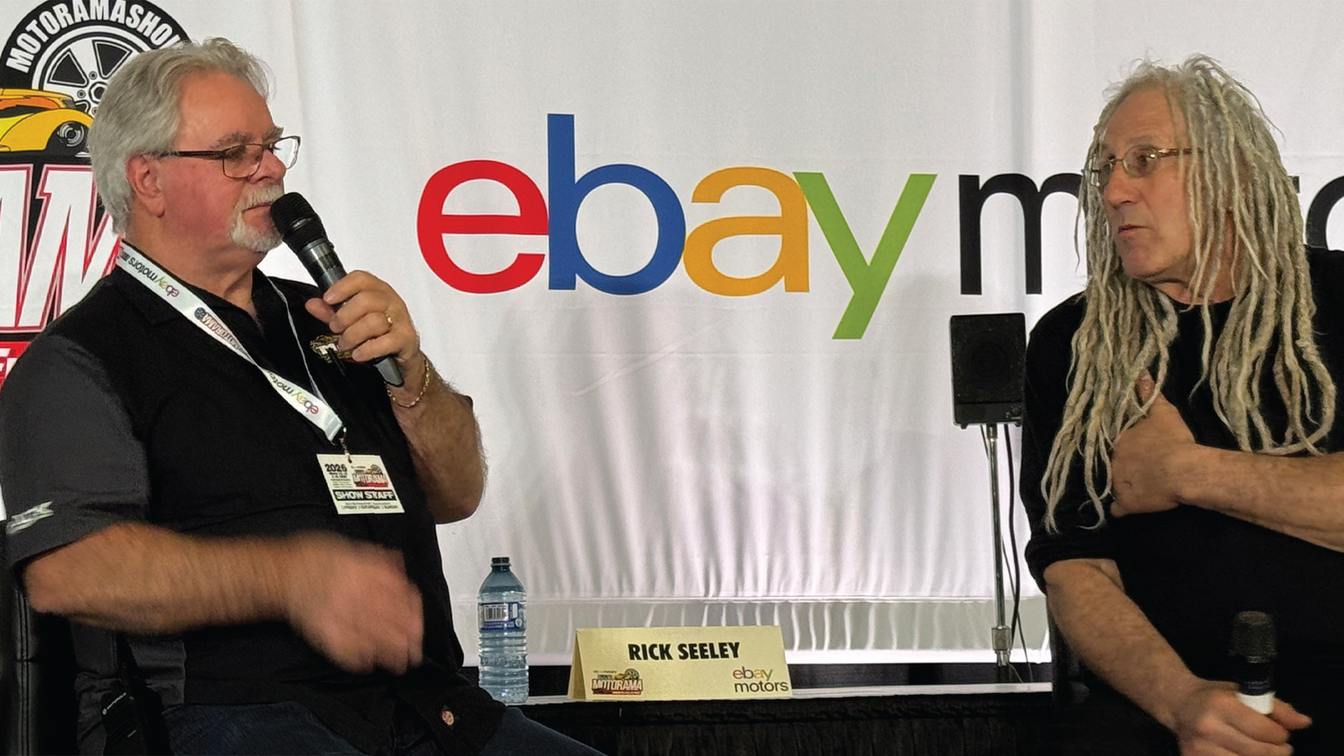 Two men speaking at a panel discussion with eBay Motors branding visible on the backdrop behind them.