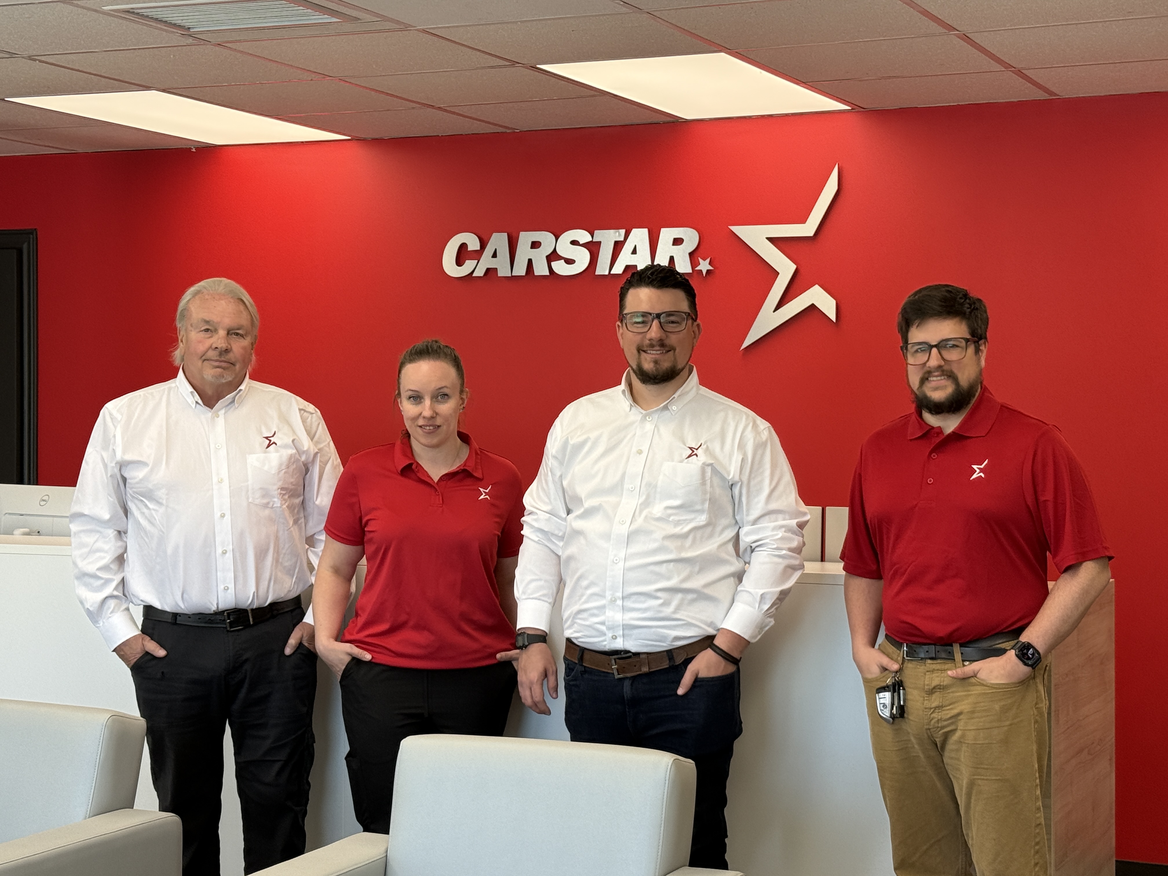 Four CARSTAR employees posing together in office with red branded wall and white star logo