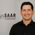 Professional headshot of smiling man in black shirt with SAAR automotive association logo background
