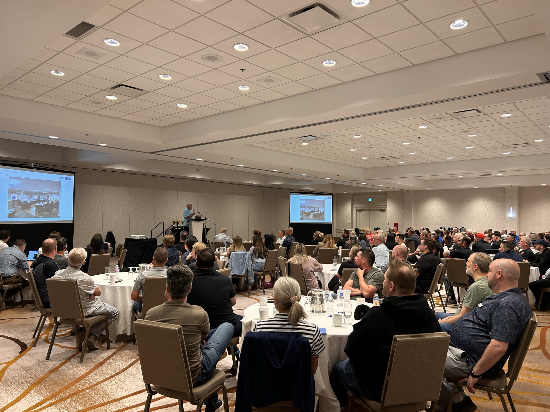 Conference attendees seated at round tables during a business presentation in a hotel ballroom with projection screens.