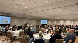 Conference attendees seated at round tables during a business presentation in a hotel ballroom with projection screens.