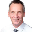 Professional headshot of smiling man in light blue shirt and dark striped tie