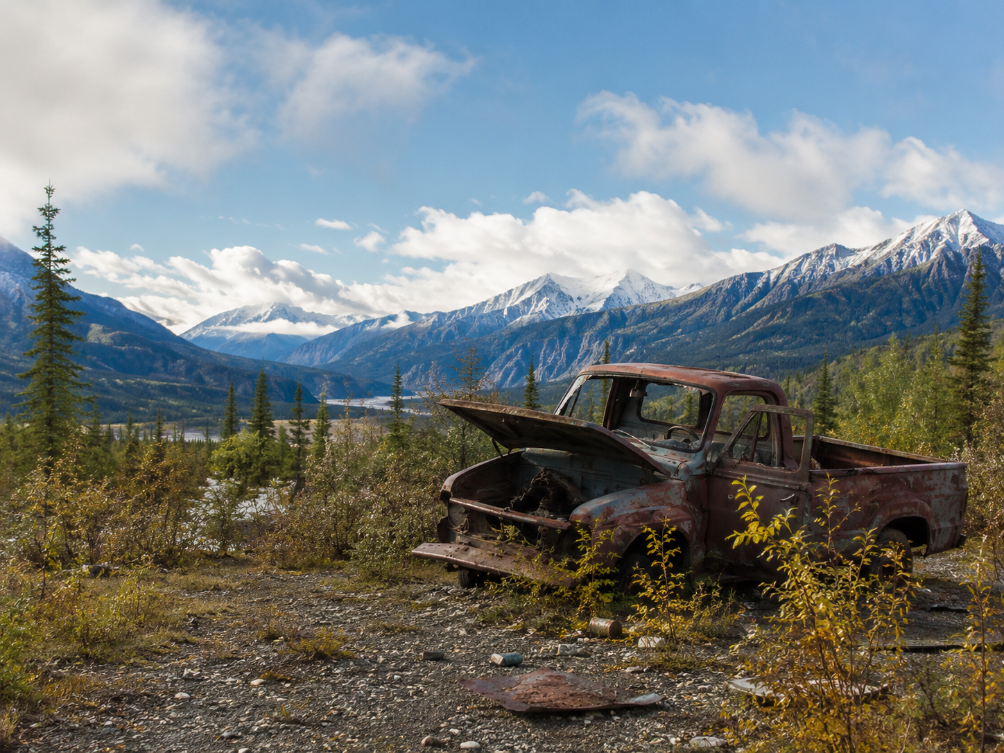 Abandoned rusted truck in alpine valley with snow-capped mountains and evergreen forest backdrop