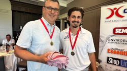 Two men wearing medals at professional event, holding fabric item with sponsor logos visible in background