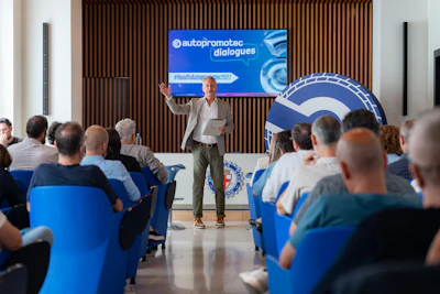 Speaker presenting at Autoproomotec Dialogues conference to seated audience in blue chairs