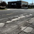Deteriorated asphalt street with multiple potholes and patched repairs near commercial buildings