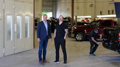 Two men in business attire stand in an automotive service facility with vehicles and equipment in the background