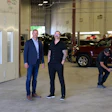 Two men in business attire stand in an automotive service facility with vehicles and equipment in the background