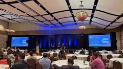 Corporate conference ballroom with attendees seated at round tables facing presentation screens with blue lighting and decorative chandeliers.