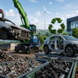 Industrial recycling facility processing vehicles with crusher equipment, metal scraps, and wind turbines in background