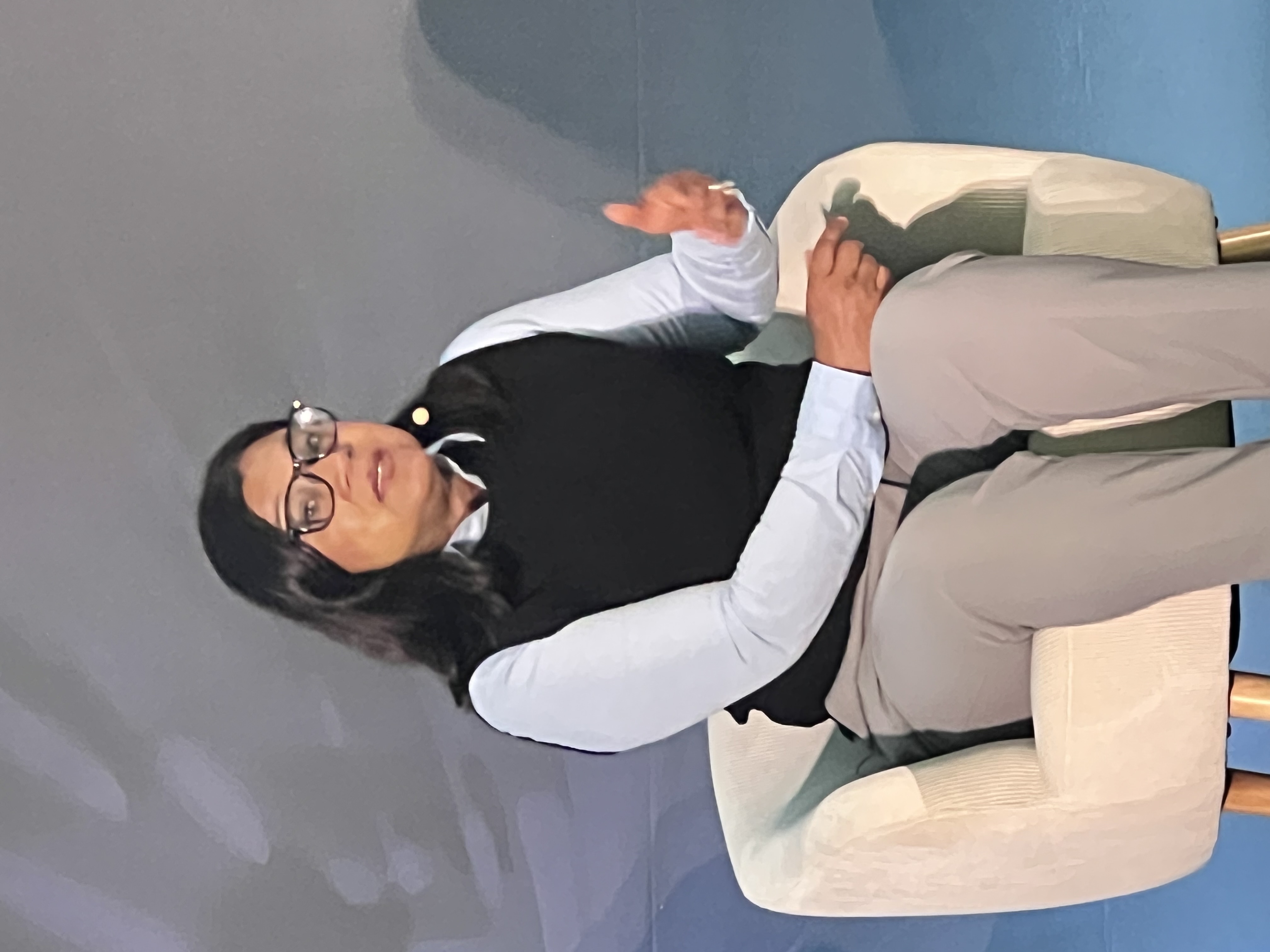 Professional woman wearing glasses and black vest speaking during a presentation while seated in a cream-colored chair