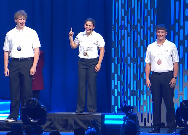 Three men in white shirts stand on stage during a professional presentation with blue backdrop and lighting.