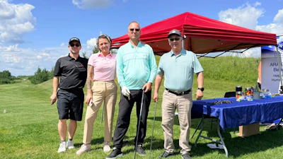 Four golfers pose together at an outdoor charity event under a red tent canopy