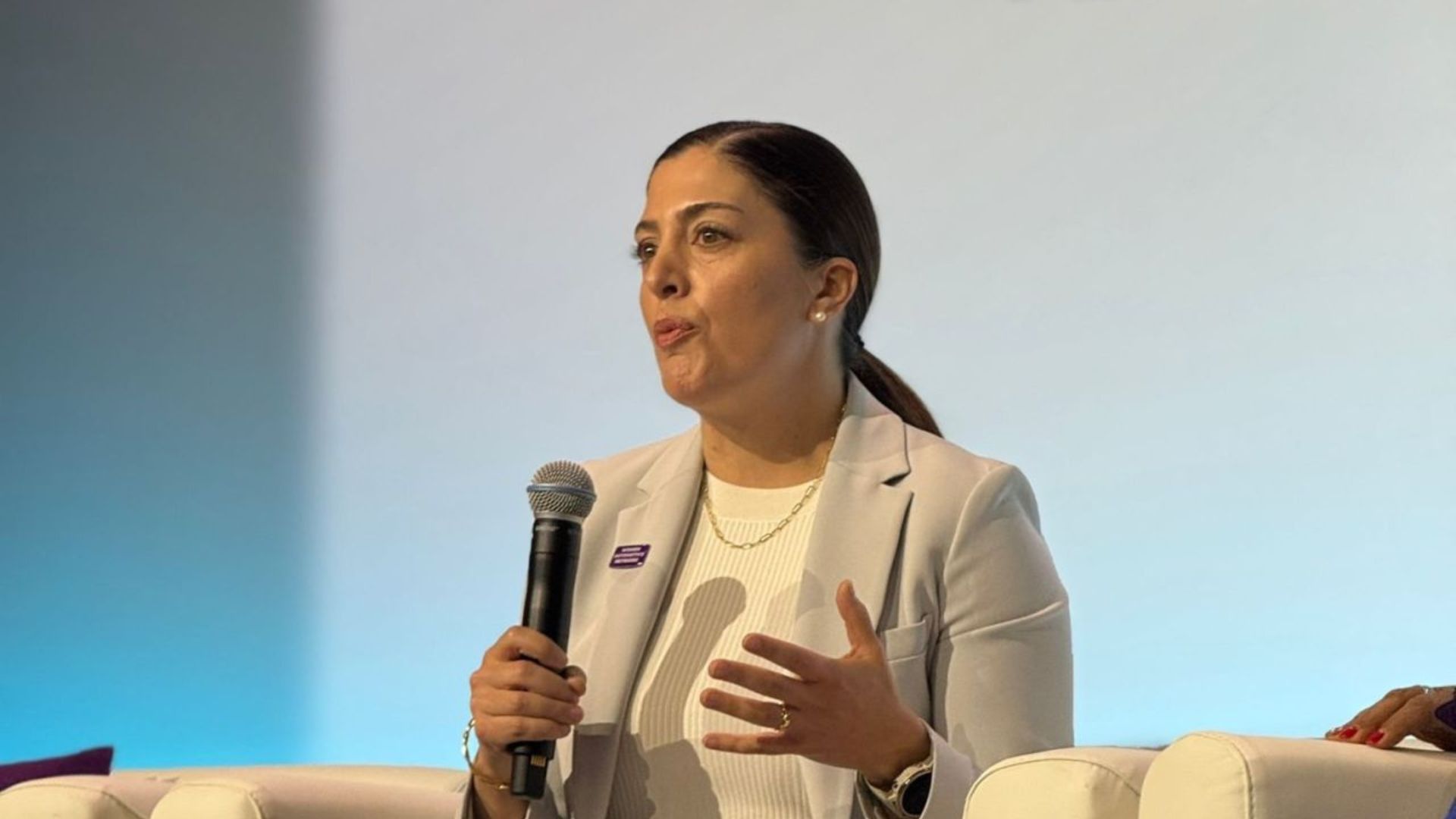 Professional woman speaking at conference panel with microphone, wearing beige blazer
