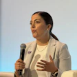 Professional woman speaking at conference panel with microphone, wearing beige blazer