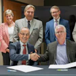 Eight business professionals pose for a formal group photo, with two men shaking hands at a table in the foreground.