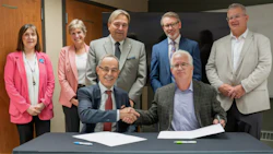 Eight business professionals pose for a formal group photo, with two men shaking hands at a table in the foreground.