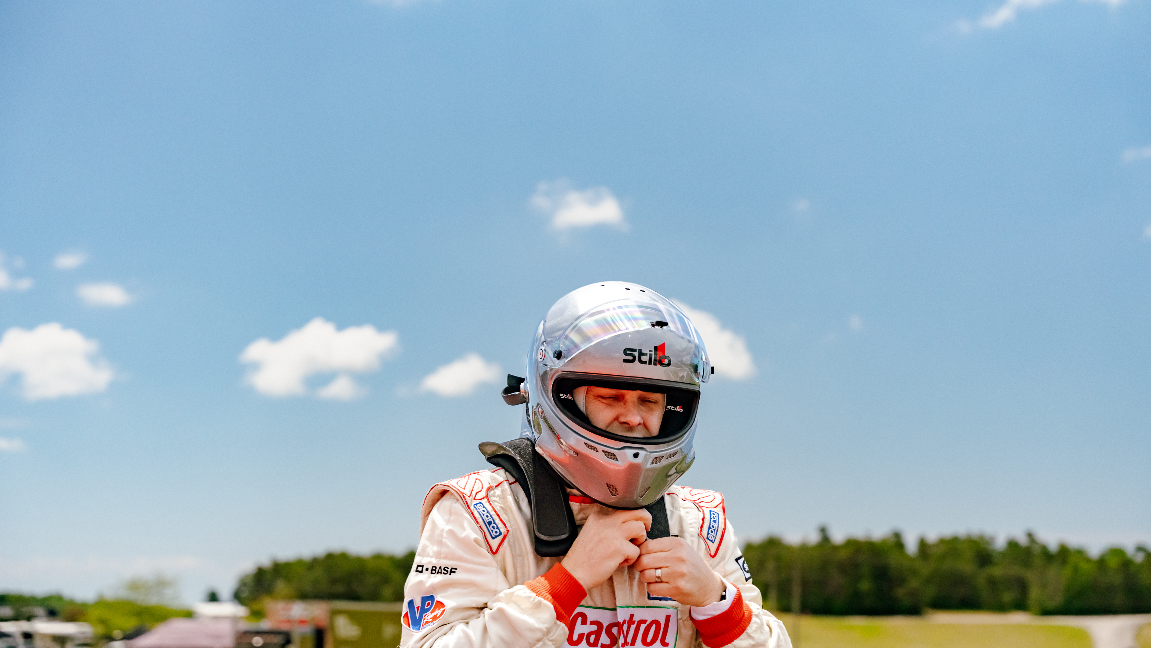 Racing driver in white Stilo helmet and red Castrol racing suit against blue sky