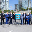 Group of officials and workers posing at gas station with price display and city buildings in background