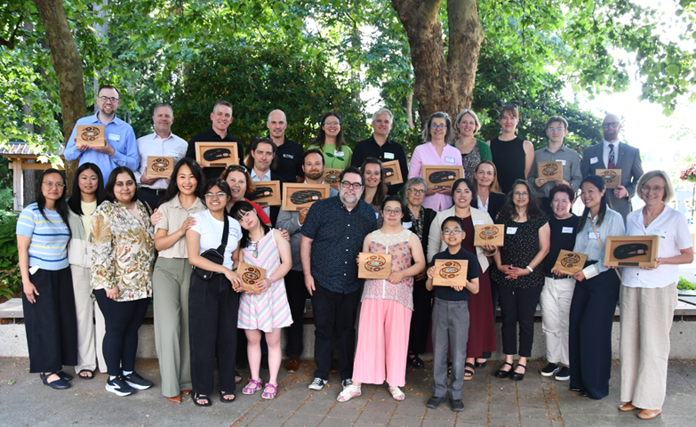 Large group of people holding wooden awards posing together outdoors under trees at an event