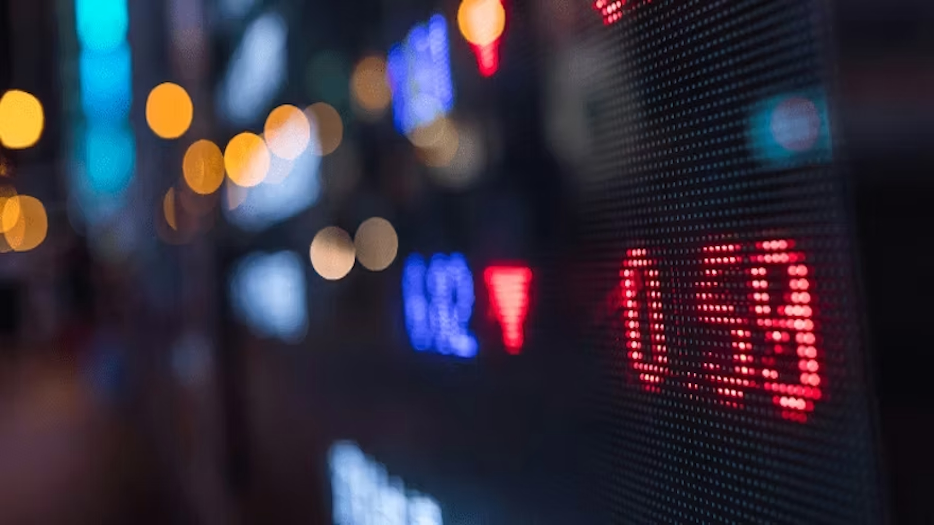 Nighttime urban street with blurred neon lights and red LED financial market display showing stock data