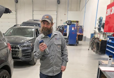 Bearded technician in gray work shirt and cap holding a tool in an automotive service garage with vehicles and equipment
