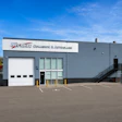 Modern industrial facility with gray exterior, white garage doors, and company signage under clear blue sky