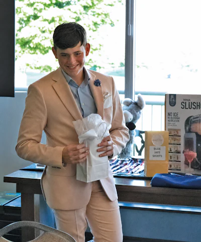 Young man in tan blazer smiling while holding white gift bag at retail event with product displays
